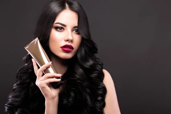 Hermosa modelo morena: rizos, maquillaje clásico y labios rojos con una botella de productos para el cabello. La cara de belleza . — Foto de Stock