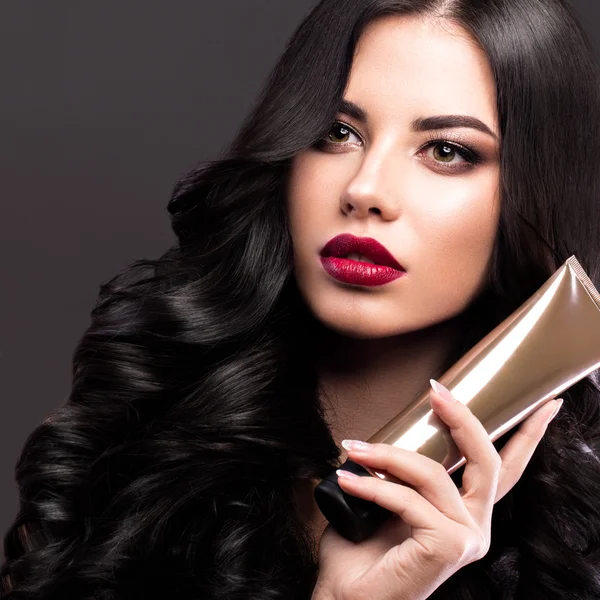 Hermosa modelo morena: rizos, maquillaje clásico y labios rojos con una botella de productos para el cabello. La cara de belleza . —  Fotos de Stock