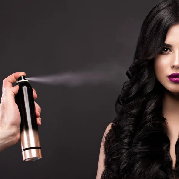 Beautiful brunette model: curls, classic makeup and red lips with a bottle of hair products. The beauty face. — Stock Photo, Image