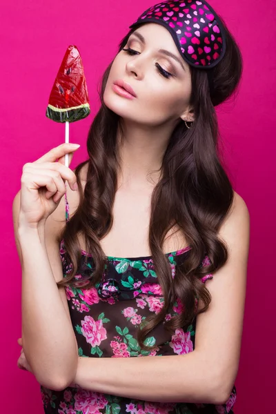 Funny young woman in sleeping mask and pajamas, sweets on pink background. beauty face. — Stock Photo, Image