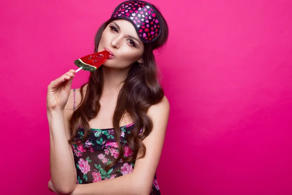 Mujer joven divertida en máscara para dormir y pijama, dulces sobre fondo rosa. cara de belleza . — Foto de Stock