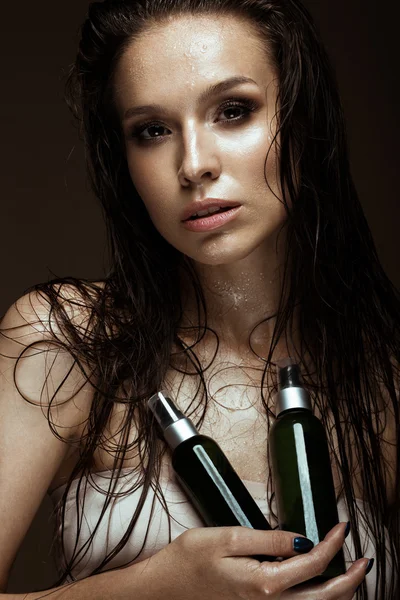 Belle fille avec un maquillage lumineux, cheveux et peau mouillés, bouteille de produits cosmétiques dans les mains. Beauté visage . — Photo