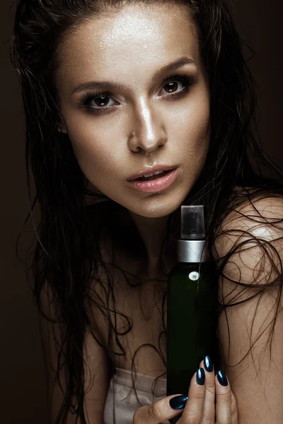 Belle fille avec un maquillage lumineux, cheveux et peau mouillés, bouteille de produits cosmétiques dans les mains. Beauté visage . — Photo