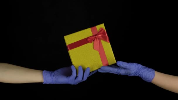 Pandemisch kerstcadeau, verrassing. COVID-19 Kerstmis 2021. Handen in blauw medische beschermende handschoenen passeren geschenkdoos met strik geïsoleerd op zwarte achtergrond — Stockvideo