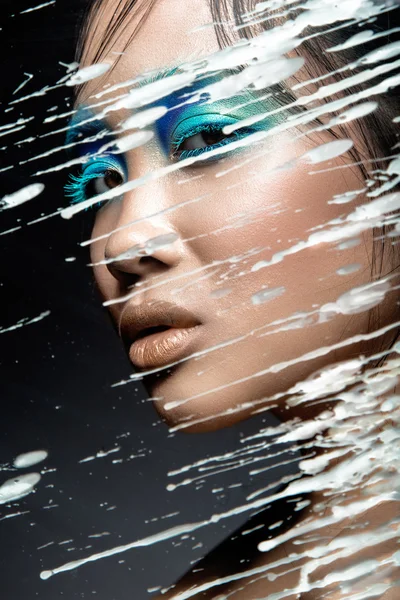 Beautiful Asian girl with bright blue make-up behind glass and drops of wax. Beauty face. — Stock Photo, Image