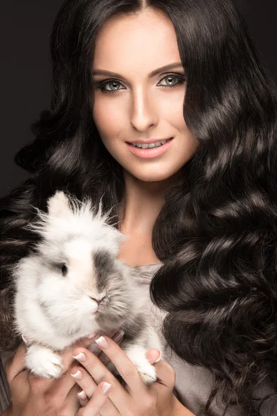Linda modelo morena menina, cachos perfeitos e maquiagem de moda à noite com um coelho em suas mãos. Cara de beleza . — Fotografia de Stock