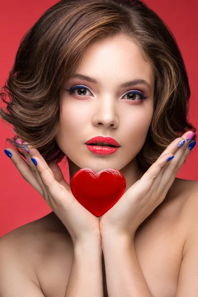 Belle fille modèle avec un maquillage lumineux et le cœur rouge dans les mains. Un beau visage. Ongles courts et colorés — Photo