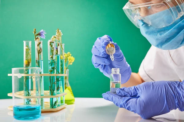Jeune femme technicienne ou scientifique charge échantillon liquide dans la bouteille avec pipette. Tubes à essai avec fleurs sur fond. — Photo