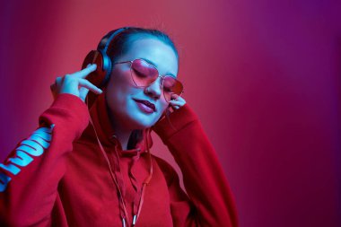 Moda hippisi kadın şık gözlükler takıyor ve kulaklık takıyor. Stüdyoda neon renkli arka planda müzik dinliyor..