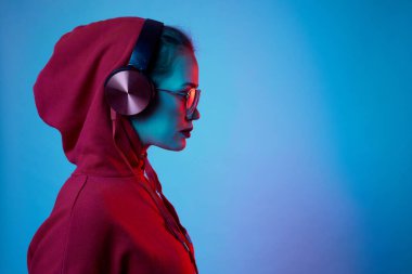 Moda hippisi kadın şık gözlükler takıyor ve kulaklık takıyor. Stüdyoda neon renkli arka planda müzik dinliyor..