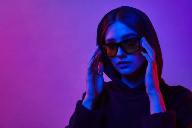 Stüdyoda neon ışığı altında poz veren bir kız. Yuvarlak güneş gözlüklü ve renkli ışık efektli bir kadın. Disko tarzı. Jel filtresi.