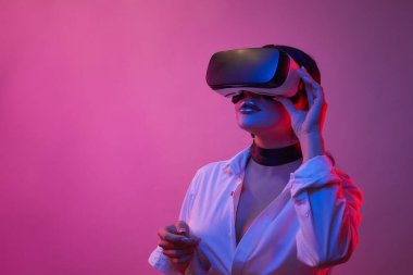 Kadın mavi arka planda neon ışıkta VR gözlükleriyle oynuyor. Sanal gerçeklik deneyimi yaşarken VR gözlüklü kız seçenekleri seçiyor.