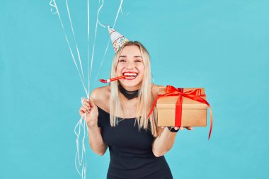 Hediye kutulu ve balonlu genç bir kadın mavi arka planda doğum gününü kutlarken kameraya bakıyor ve parti kornası çalıyor.