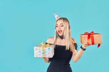 Siyah elbiseli ve doğum günü şapkalı güzel bir kadın mavi arka planda hediye kutusu tutuyor. Sevimli mutlu genç kız doğum günü partisini kutluyor..