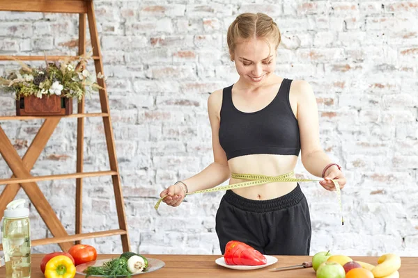 Femeia tânără pe o dietă, își măsoară talia cu o bandă moale de măsurare, legume proaspete pe masă — Fotografie, imagine de stoc