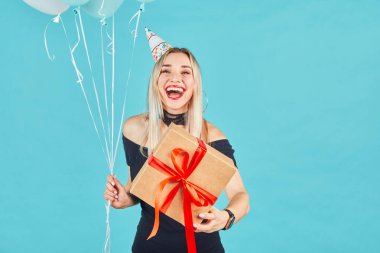 Siyah elbiseli ve doğum günü şapkalı güzel bir kadın elinde hediye kutusu ve mavi arka planda pastel hava balonları tutuyor. Mutlu genç kız doğum günü partisini kutluyor.