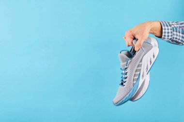 Mavi pastel arka planda gri spor ayakkabıları asılı duruyor. Yeni bir spor ayakkabıyla el. Stabilite ve minder koşu ayakkabıları. Kapat..