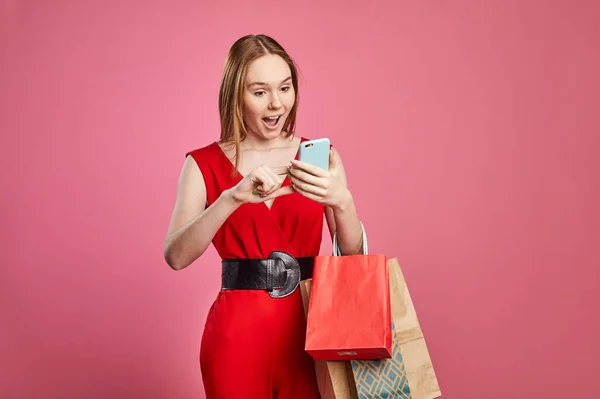 Estúdio tiro de mulher feliz comprador segurando sacos de compras usando aplicativos móveis para compras on-line em fundo rosa. Comércio eletrônico, venda, descontos de shopping no conceito de aplicações. — Fotografia de Stock