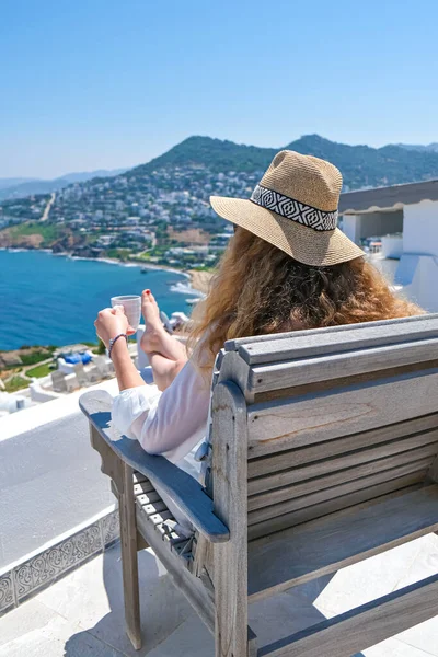 美丽的年轻女子草帽和咖啡杯坐在有海景的房子或酒店的白色阳台上 — 图库照片
