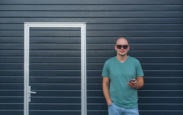 Jonge man in zonnebril met telefoon tegen de achtergrond van een muur en de gesloten deuren — Stockfoto