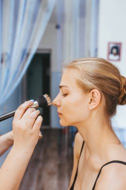 Makyaj sanatçısı, modelin yüzünde fırçayla çalışıyor. Bir güzellik salonunun içinde genç bir kadının portresi. Yan görünüm