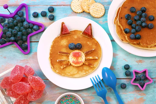 Roliga pannkakor för barn. Katt formad pannkaka på vit platta, blå trä bakgrund, ovanifrån. Kreativ idé för barn frukost. Stockfoto