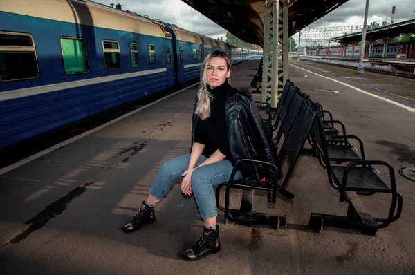 Tren istasyonundaki trenin yanında deri ceketle bankta oturan güzel bir kadının portresi. St. Petersburg 'daki Vitebsk tren istasyonunu bekliyorum..