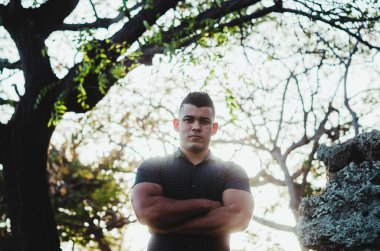 Beyaz tenli erkek modeli Venezuela kökenli Dominik Cumhuriyeti 'nden. Parkta kotlu seksi bir adam turuncu tenli, güneş ışığında yürür yaprakların arasında kollarını kavuşturur.