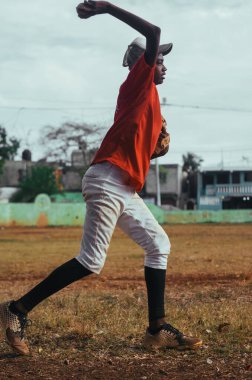 Kahverengi tenli ve latin kökenli çocuk parkta top, eldiven ve sopayla beyzbol oynuyor. Siyah Dominikli çocuk beyzbol üniforması ve şapkası var.