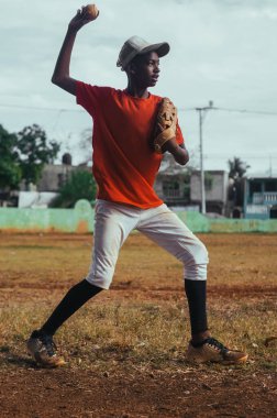 Kahverengi tenli ve latin kökenli çocuk parkta top, eldiven ve sopayla beyzbol oynuyor. Siyah Dominikli çocuk beyzbol üniforması ve şapkası var.