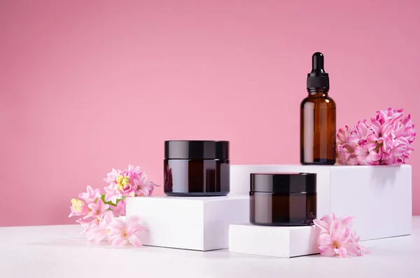 Packaging for cosmetic cream and essential oil of amber glass mockup on white podiums with fresh spring hyacinth flowers on pink background.