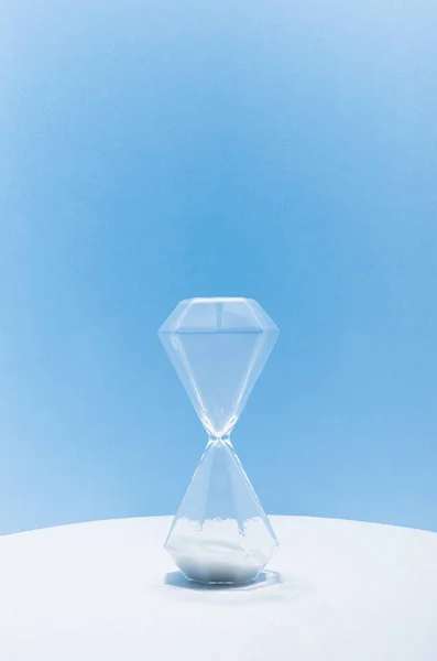 Transparent hourglass with white sand on white wood table, soft light blue wall, vertical, copy space. Time is end.
