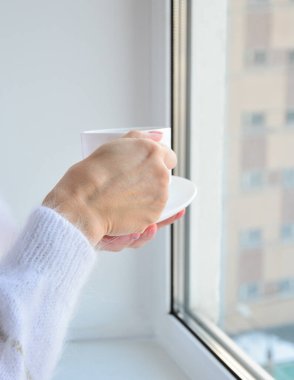 Beyaz kazaklı kadın elleri pencerenin önünde bir fincan kahve tutuyor. Boşluğu kopyala