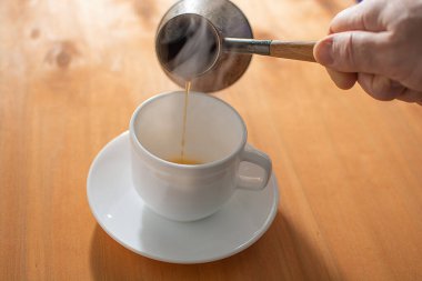 view of hot coffee being poured from a turk into a white cup, wooden background. steam from hot coffee. copy space