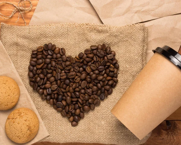 heart shape made up of coffee beans on burlap. a cardboard cup for coffee and a cookie. wooden background. the concept of dating and meeting, love and adventure. flat lay.