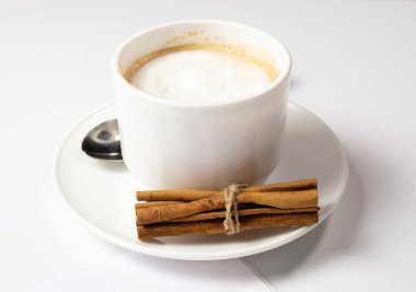 Sütlü köpüklü beyaz bir fincan kahve ve çay tabağında tarçın çubukları beyaz bir masa üzerinde beyaz bir kağıt levha ile duruyor. Boşluğu kopyala.