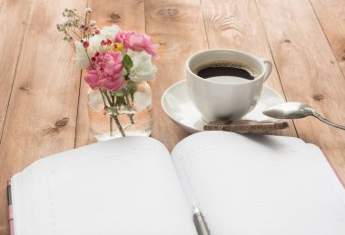 Küçük bir cam su kavanozunda pembe ve beyaz çiçeklerin olduğu bir işyeri manzarası, ayrıca ahşap bir masanın üzerinde bir kalem ve kahve bulunan kırmızı bir defter. Boşluğu kopyala.