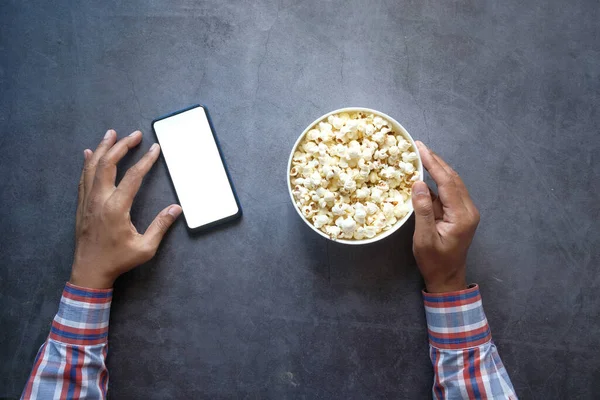 Mano del hombre usando el teléfono inteligente y comer palomitas de maíz —  Fotos de Stock