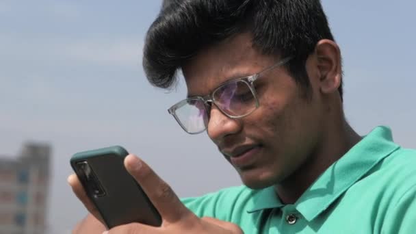 Усміхнений молодий чоловік в окулярах використовує свій телефон на відкритому повітрі — стокове відео
