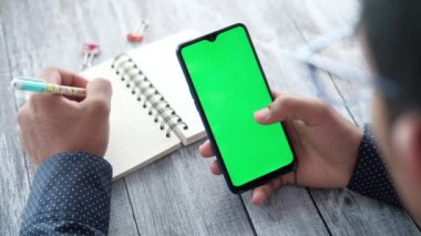 Akıllı telefon kullanan ve not defterine yazı yazan genç bir adamın arka penceresi 