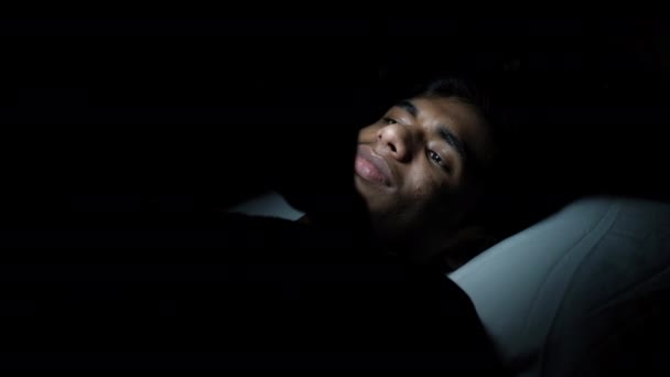 Joven sentado en la cama usando un teléfono inteligente por la noche — Vídeo de stock