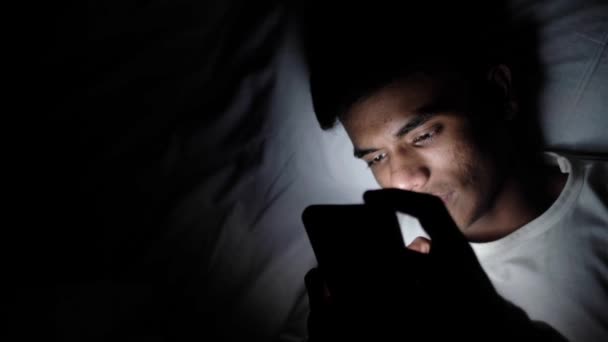 Genç adam geceleri akıllı telefon kullanarak yatakta oturuyor. — Stok video