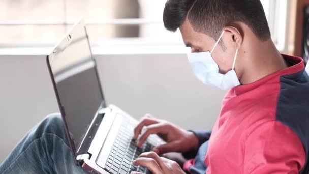 Vista lateral del hombre joven en máscara quirúrgica utilizando el ordenador portátil en casa — Vídeo de stock