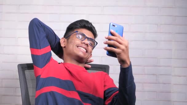 Glad ung asiatisk man sitter på stol med hjälp av smarttelefon. — Stockvideo