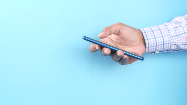 Visão lateral da mão jovem usando telefone inteligente contra fundo azul — Vídeo de Stock