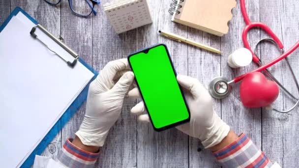 Oversikten over legens hånd i latekshansker som holder smarttelefon med grønn skjerm – stockvideo