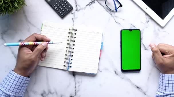 Bovenaanzicht van de hand van de mens met behulp van smart phone en schrijven op notitieblok — Stockvideo