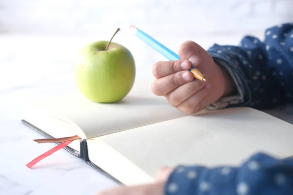 Kind meisje hand schrijven op notitieblok met groene verse appel op tafel — Stockfoto