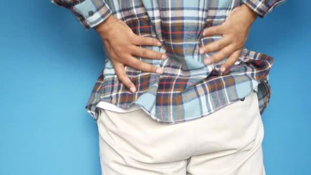 Молодой человек страдает от боли в спине, стоя на синем фоне — стоковое видео