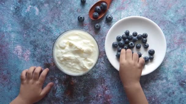 Детская рука кладет голубую ягоду на свежий йогурт — стоковое видео